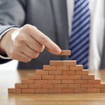 Businessman in a suit putting the last piece of a pyramid using wooden blocks - success concept